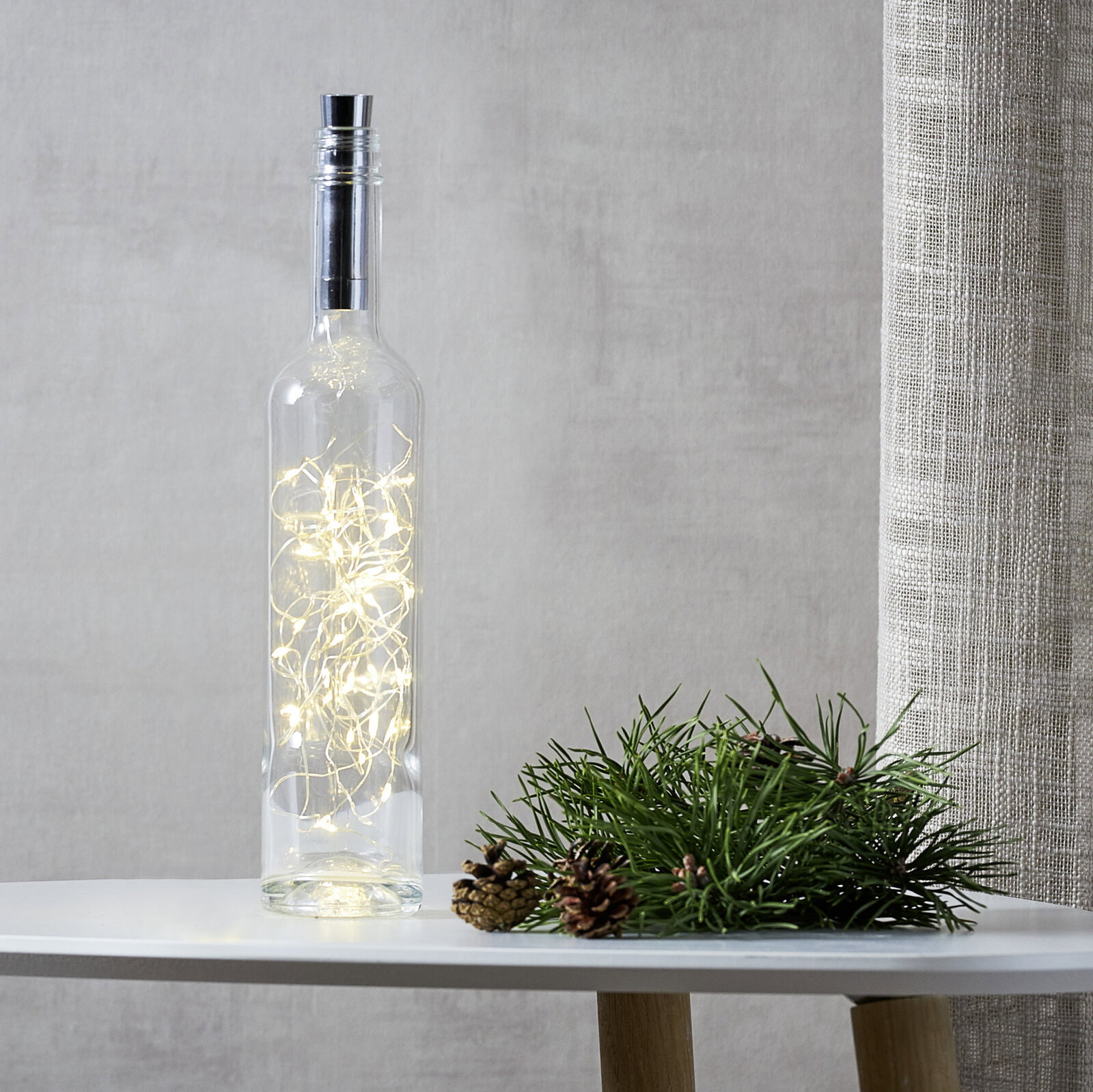 Transparente Glasflasche mit warmweiß leuchtender LED-Lichterkette im Inneren, auf einem weißen Tisch neben Tannenzweig und Zapfen, frontal aus leicht seitlicher Perspektive aufgenommen.