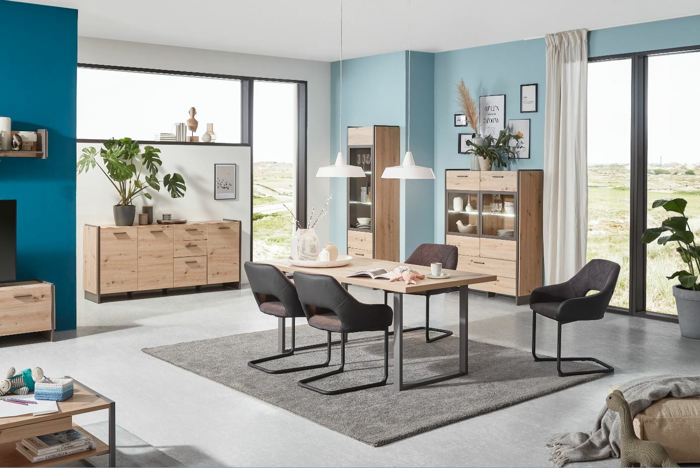 Moderne Esszimmermöbel in einem hellen Raum mit großen Fenstern, bestehend aus einem Esstisch mit vier Stühlen, einer Vitrine und einer Kommode, aus hellem Holz mit schwarzen Akzenten. Perspektive: Frontalansicht.