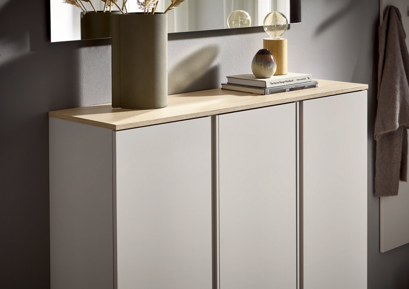 Modernes Sideboard mit drei weißen Fronten und heller Holzplatte, dekoriert mit Vase, Büchern und Lampe, in schräger Frontansicht von links.