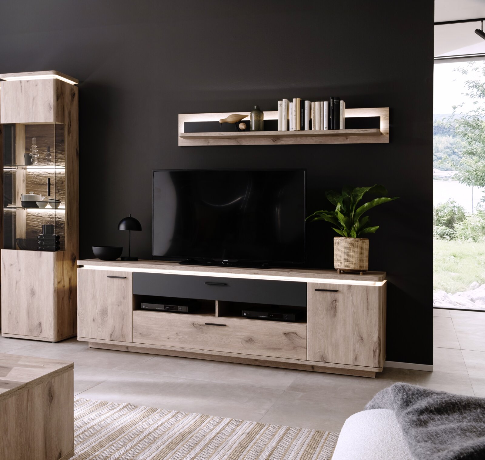 Moderne woonkamerinrichting met een tv-lowboard van licht hout en een zwart front, een bijpassende wandplank en een vitrinekast met ledverlichting tegen een donkere muur, gefotografeerd vanuit een licht schuin perspectief.
