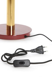 Nahaufnahme einer Tischleuchte mit rotem Sockel und goldfarbenem Ständer, inklusive schwarzem Kabel mit Schalter und Stecker.