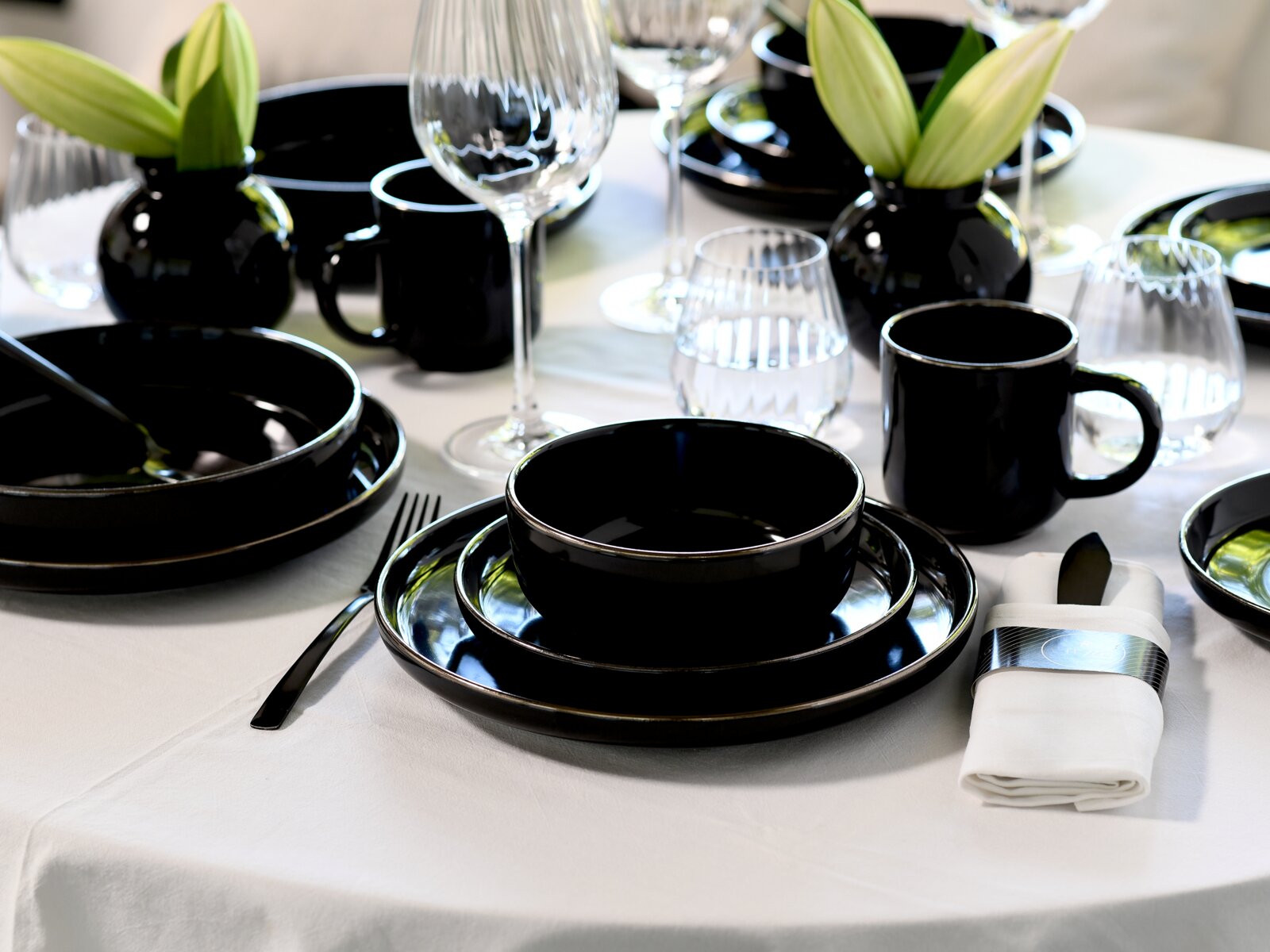 Elegantes schwarzes Geschirrset mit Schalen, Tellern und Bechern auf weißer Tischdecke, ergänzt durch Kristallgläser und kleine Vasen mit grünen Blättern, aufgenommen aus leicht erhöhter schräger Perspektive