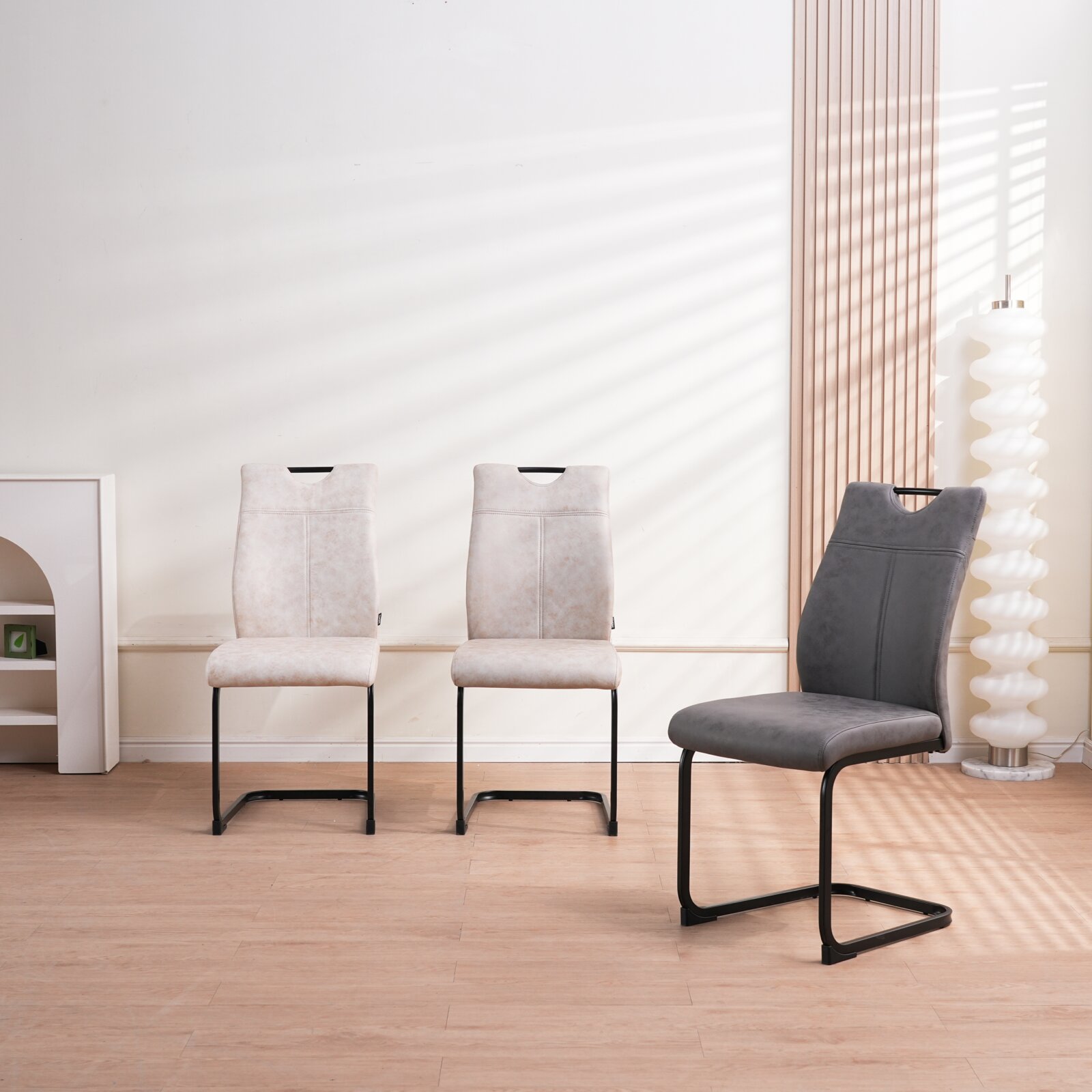 Moderne eetkamerstoel met grijze, zacht aanvoelende bekleding en zwart metalen sledeframe, gefotografeerd schuin van voren in een lichte woonkamer; op de achtergrond staan twee lichtbeige stoelen.