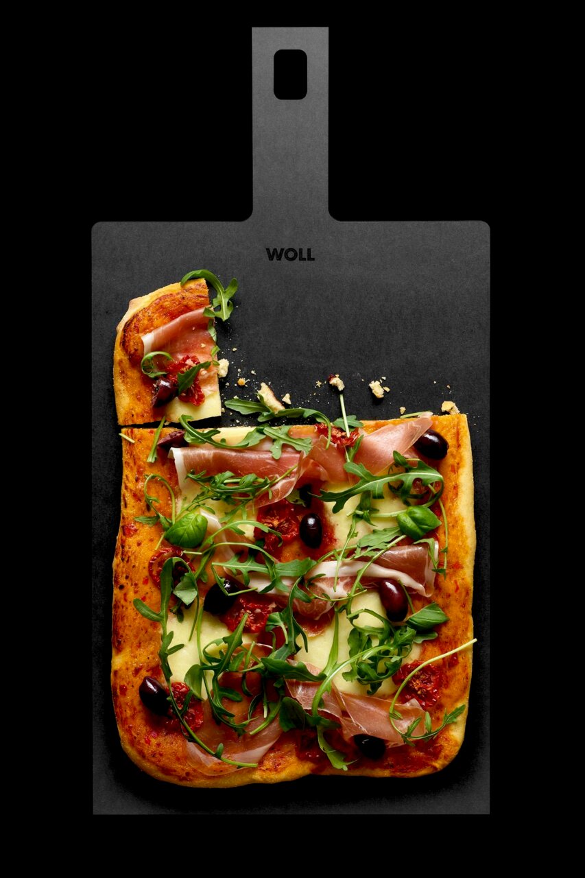 Schneidebrett mit Griff, darauf eine Pizza mit Schinken, Rucola und Oliven, aus der Vogelperspektive fotografiert.