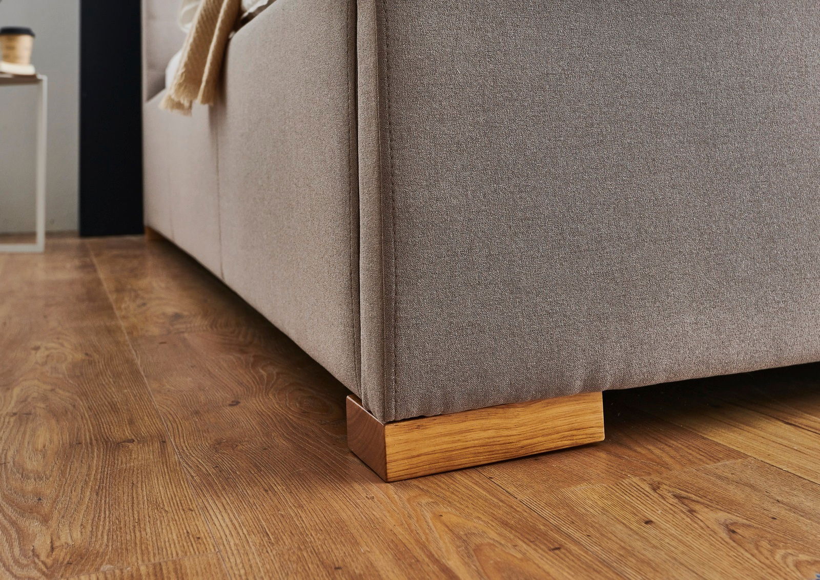 Nahaufnahme eines Polsterbetts mit hellbraunem Stoffbezug und Holzfuß auf einem Holzboden, seitliche Perspektive.