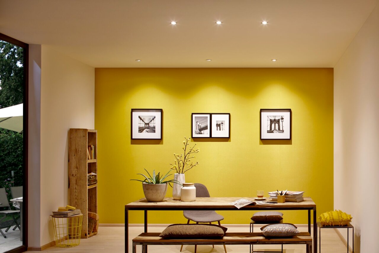 Moderne Innenraumbeleuchtung mit LED-Leuchtmitteln, die von der Decke auf einen stilvoll eingerichteten Raum mit gelber Akzentwand und Holzmöbeln strahlt. Perspektive von vorne.