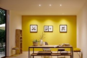 Moderne Innenraumbeleuchtung mit LED-Leuchtmitteln, die von der Decke auf einen stilvoll eingerichteten Raum mit gelber Akzentwand und Holzmöbeln strahlt. Perspektive von vorne.