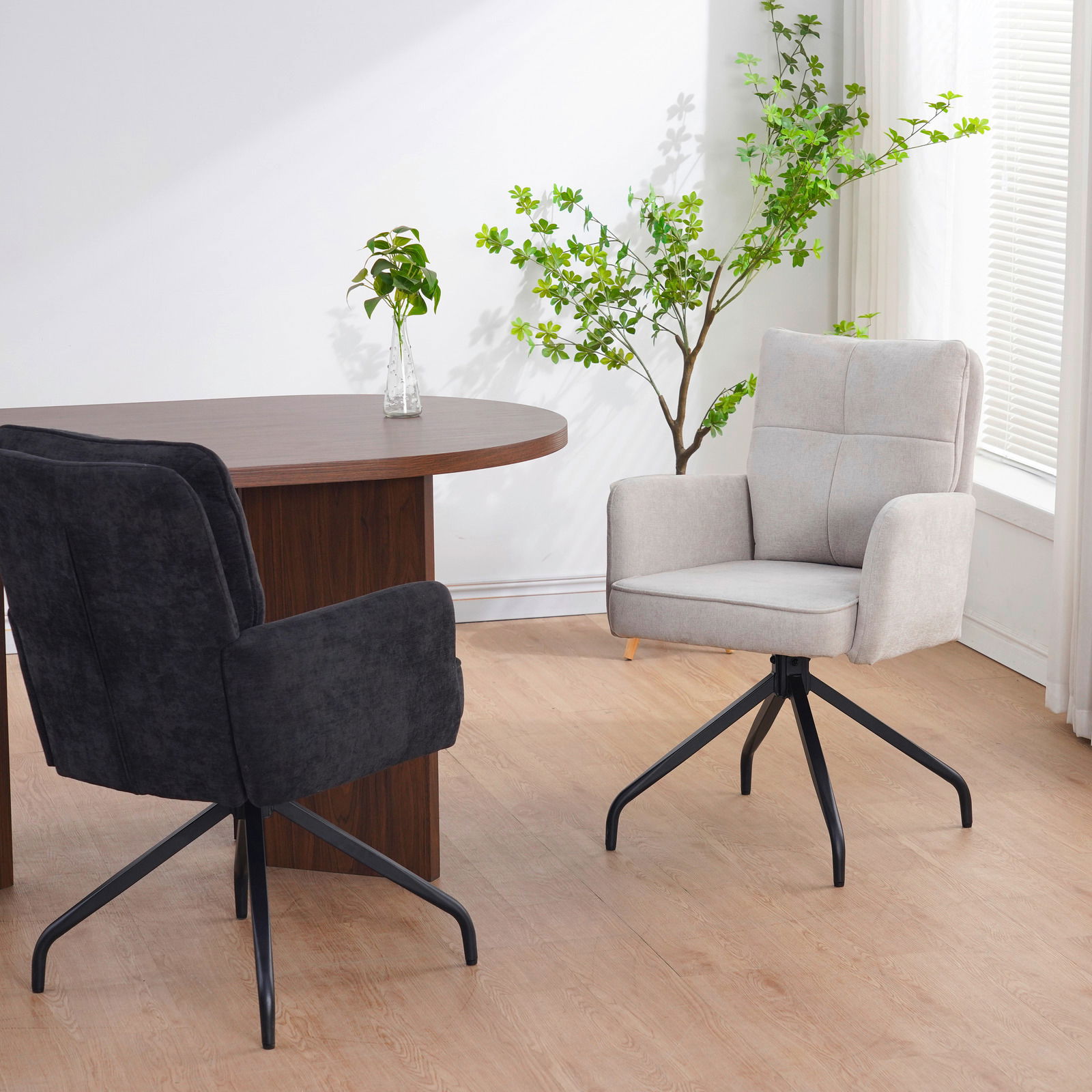 Zwei Schwingstühle in einem modernen Raum, einer in hellem Stoff und der andere in dunklem Stoff, mit einem runden Holztisch und Pflanzen im Hintergrund. Perspektive von der Seite.