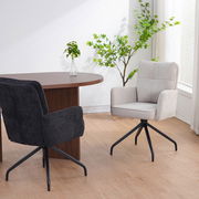 Zwei Schwingstühle in einem modernen Raum, einer in hellem Stoff und der andere in dunklem Stoff, mit einem runden Holztisch und Pflanzen im Hintergrund. Perspektive von der Seite.