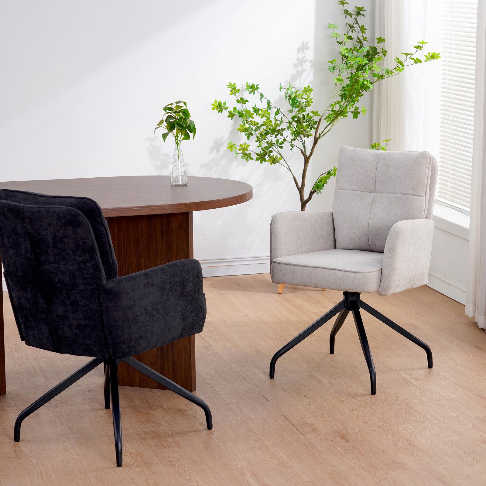 Zwei Schwingstühle in einem modernen Raum, einer in hellem Stoff und der andere in dunklem Stoff, mit einem runden Holztisch und Pflanzen im Hintergrund. Perspektive von der Seite.