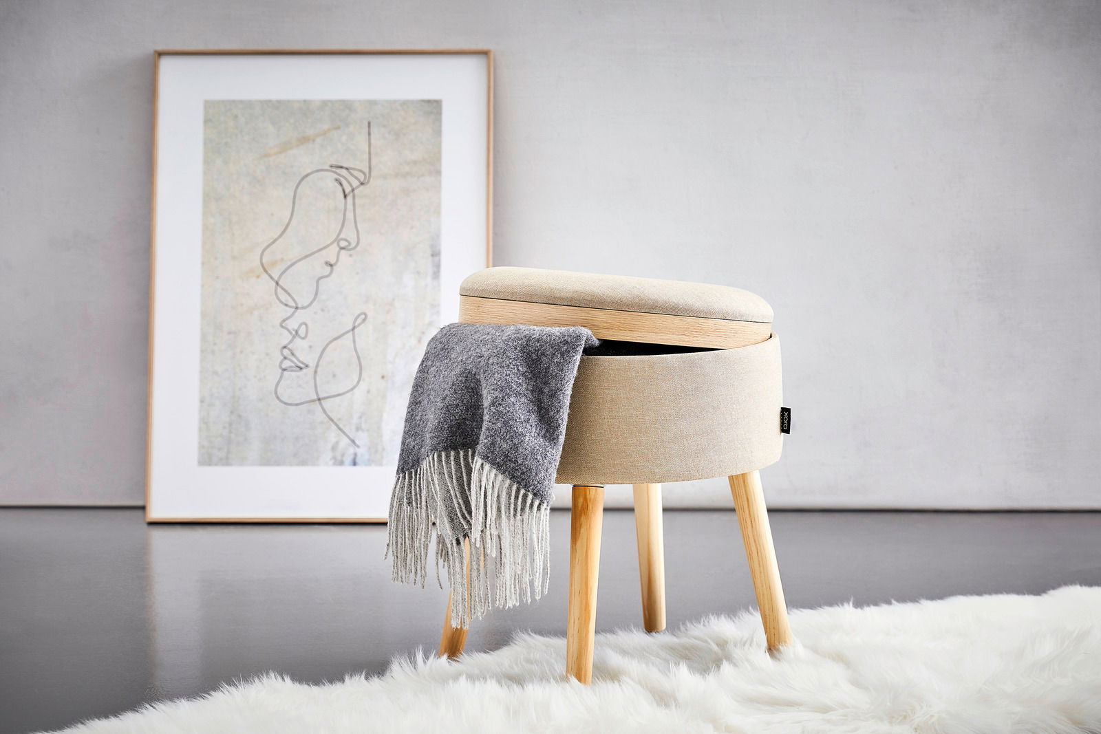 Moderner Hocker mit Stauraum, beige gepolstert, auf Holzbeinen, seitliche Perspektive, mit grauer Decke und Kunstwerk im Hintergrund.