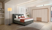 Sfeervolle slaapkamer met grijs gestoffeerd boxspringbed met hoog hoofdbord, witte matrassen en roestkleurige sprei, naast een staande lamp en een beige houten kledingkast; perspectief: schuin van voren (3/4-aanzicht)