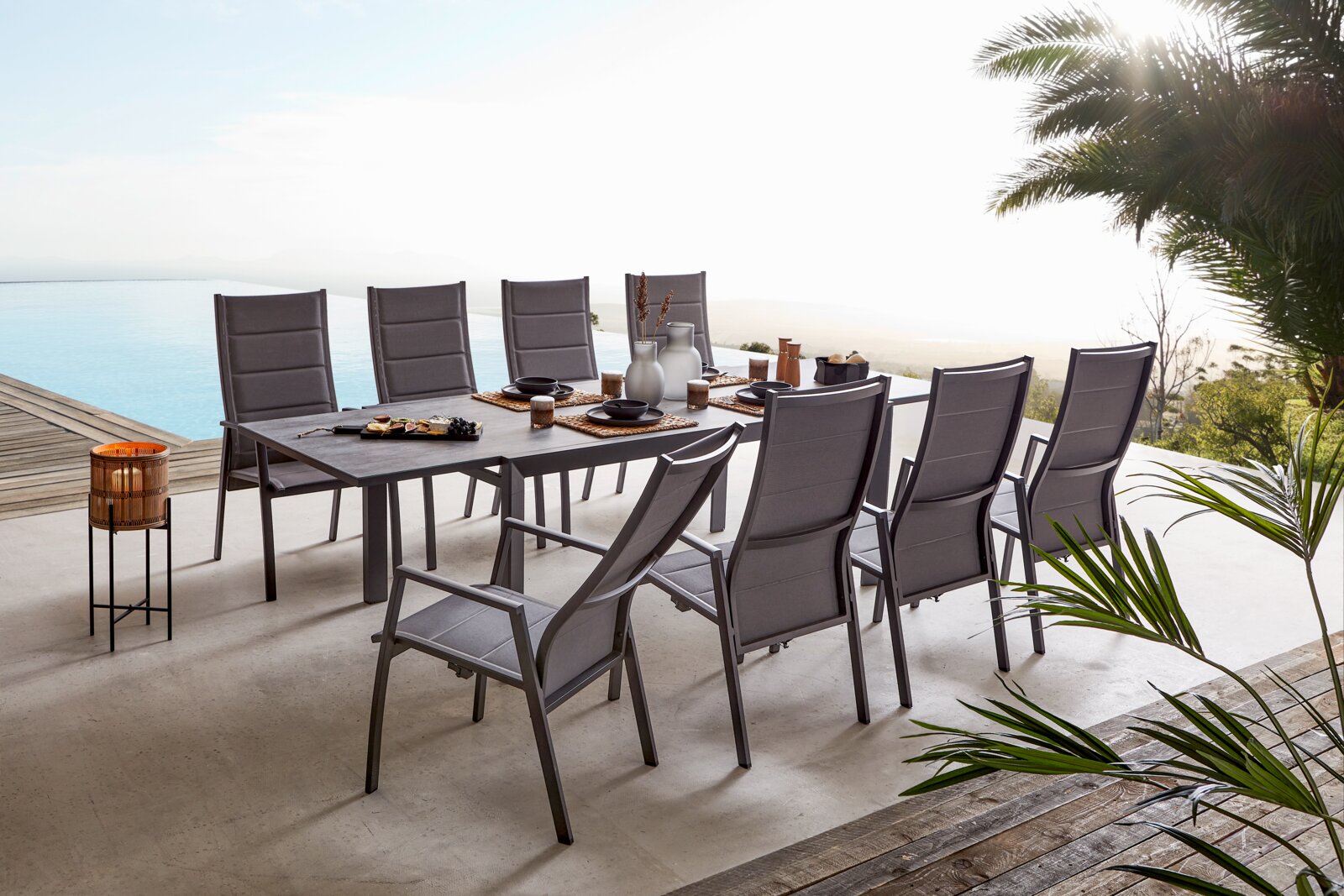 Modernes Garten-Ess-Set mit großem rechteckigem Tisch und acht grauen Hochlehner-Stühlen auf einer Terrasse am Pool, gedeckt mit Geschirr; Perspektive: leicht erhöhte schräge Seitenansicht von vorn rechts.