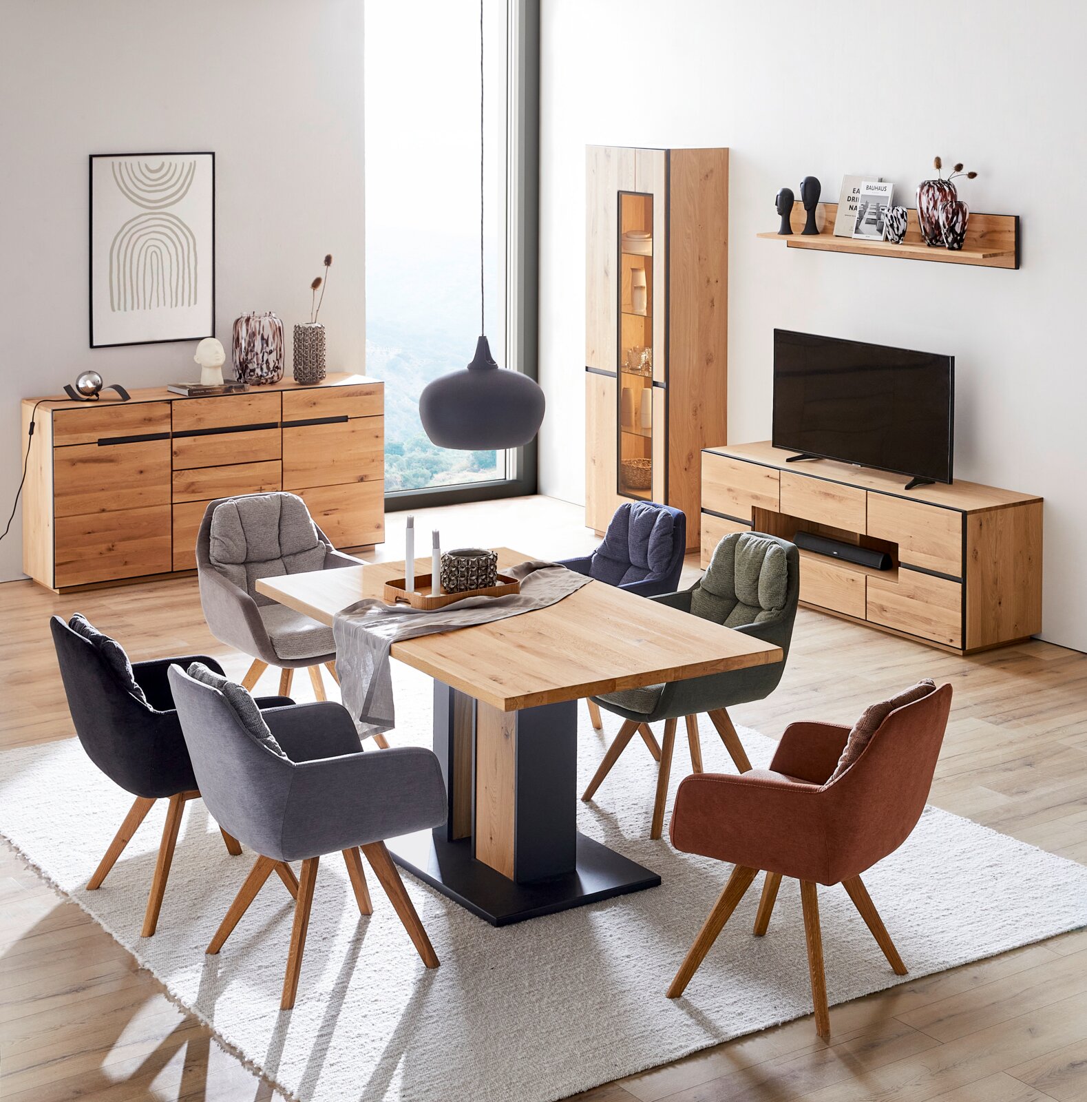 Schuine vooraanzicht van een modern eet- en woonkamerinterieur met eikenhouten eettafel, gestoffeerde stoelen in verschillende kleuren, houten dressoir en tv-meubel, vitrinekast en wandplank, met grote raampartij en zwarte hanglamp.