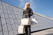Frau in schwarzer Jacke hält eine sandfarbene Shopper-Tasche mit Herringbone-Muster vor moderner Architektur, seitliche Perspektive.