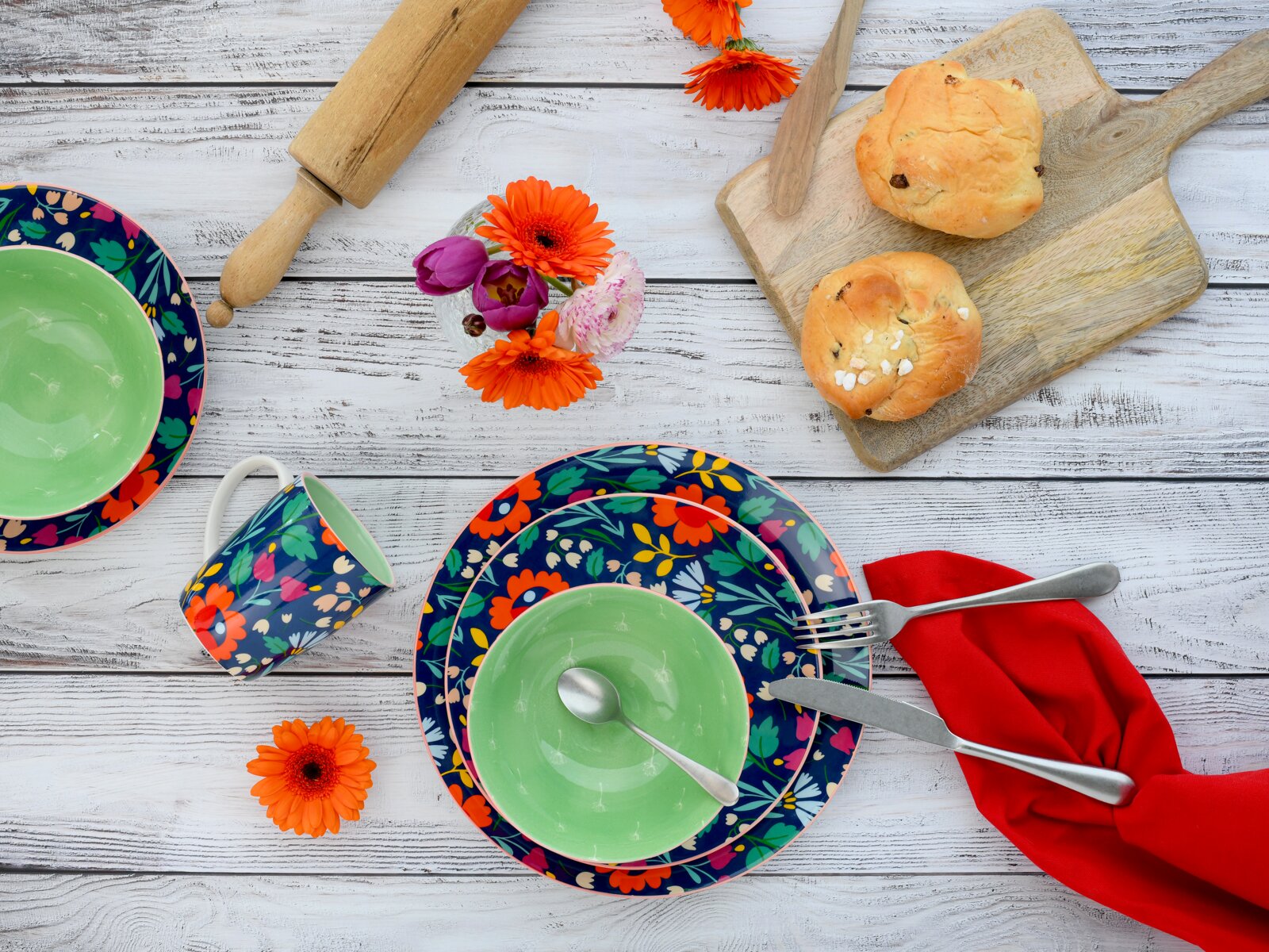 Buntes Frühstücks-Geschirrset mit floralen Tellern, grüner Schüssel und Tasse auf weißer Holzoberfläche, daneben Brötchen auf Holzbrett, Besteck und rote Serviette, Draufsicht von oben