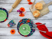Buntes Frühstücks-Geschirrset mit floralen Tellern, grüner Schüssel und Tasse auf weißer Holzoberfläche, daneben Brötchen auf Holzbrett, Besteck und rote Serviette, Draufsicht von oben