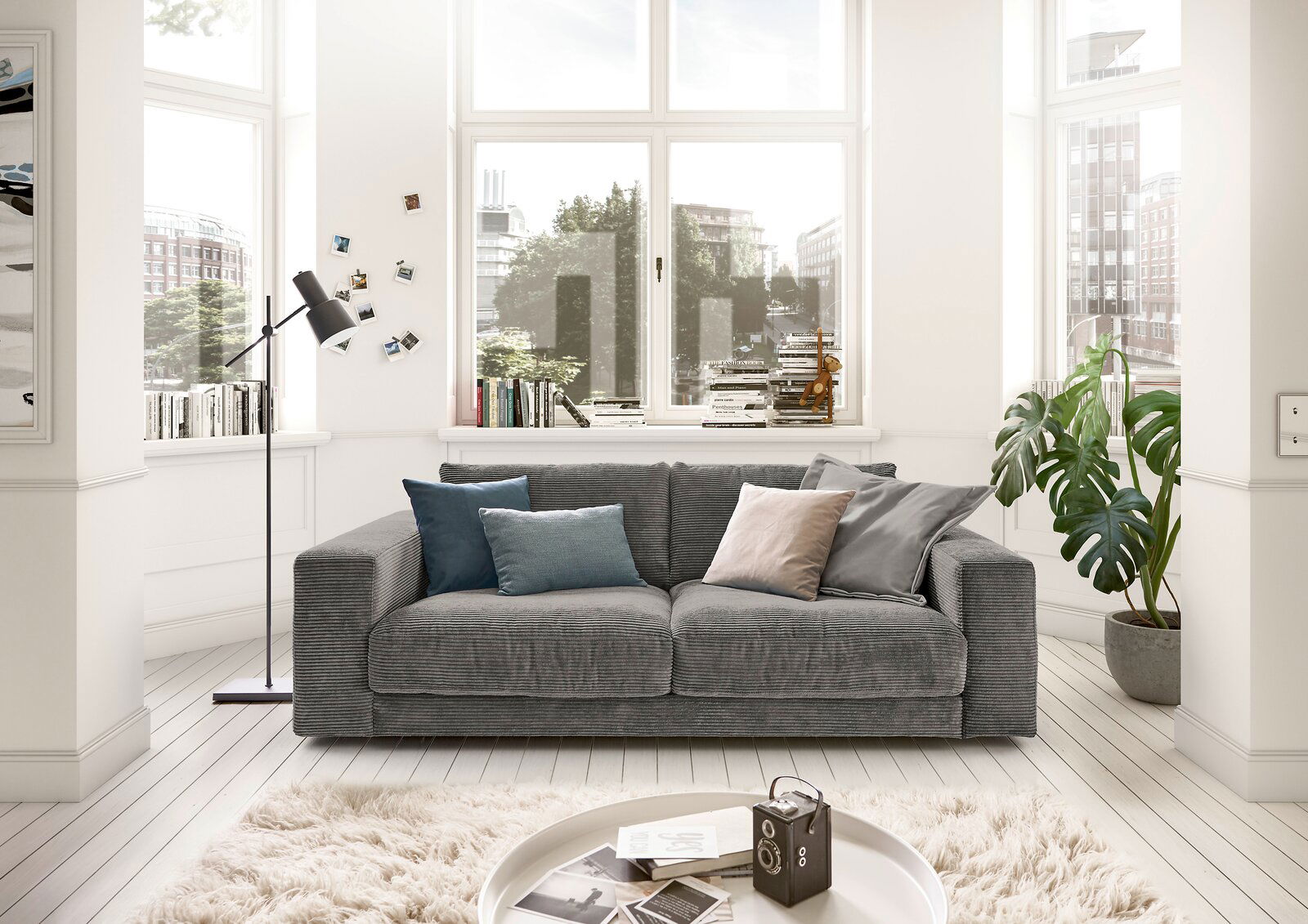 Graues 2-Sitzer-Sofa in einem hellen Wohnzimmer mit großen Fenstern, dekoriert mit bunten Kissen. Frontalansicht.