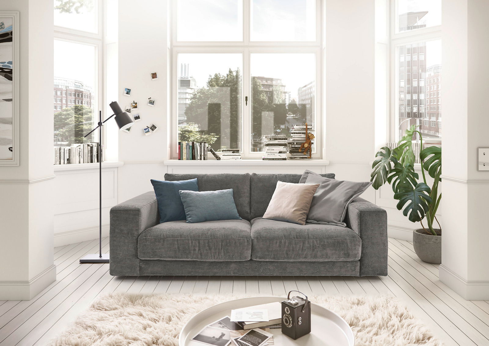 Graues 2-Sitzer-Sofa in einem hellen Wohnzimmer mit großen Fenstern, dekoriert mit bunten Kissen. Frontalansicht.