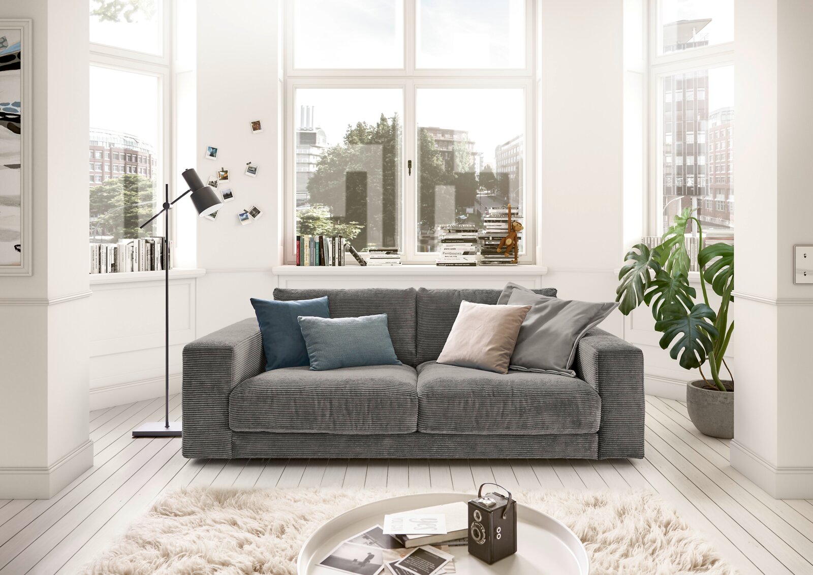 Graues 2-Sitzer-Sofa in einem hellen Wohnzimmer mit großen Fenstern, dekoriert mit bunten Kissen. Frontalansicht.