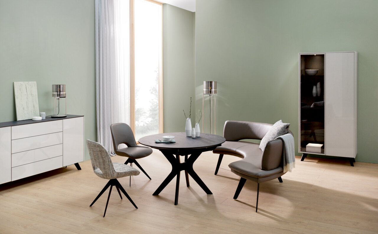 Moderne Wohnzimmermöbel in einem hellen Raum mit grün gestrichenen Wänden, bestehend aus einem runden Esstisch, zwei Stühlen und einer geschwungenen Sitzbank, aus seitlicher Perspektive betrachtet.