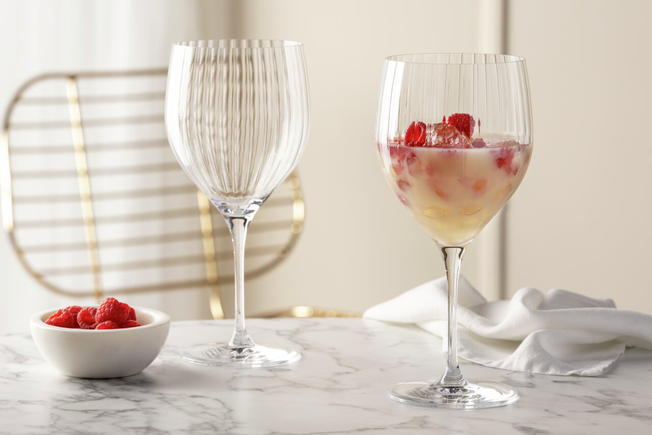 Zwei elegante Cocktailgläser POESIA 750ml auf einem Marmortisch, eines gefüllt mit einem fruchtigen Getränk und Himbeeren, im Hintergrund ein stilvoller Stuhl. Perspektive: seitlich.