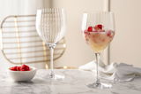 Zwei elegante Cocktailgläser POESIA 750ml auf einem Marmortisch, eines gefüllt mit einem fruchtigen Getränk und Himbeeren, im Hintergrund ein stilvoller Stuhl. Perspektive: seitlich.