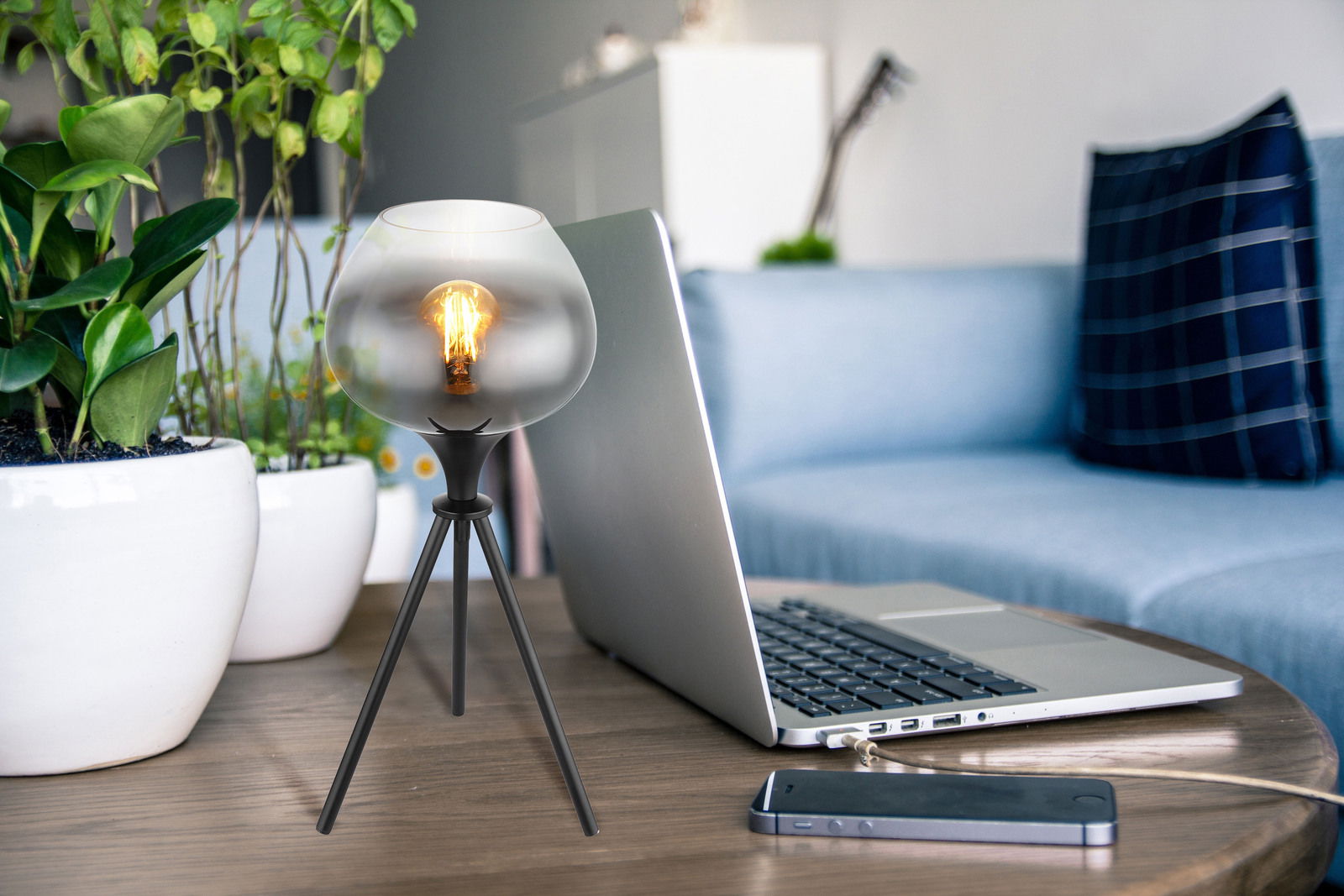 Moderne Tischleuchte mit dreibeinigem Stativ und kugelförmigem Glaslampenschirm, auf einem Tisch neben einem Laptop und Pflanzen platziert, seitliche Perspektive.
