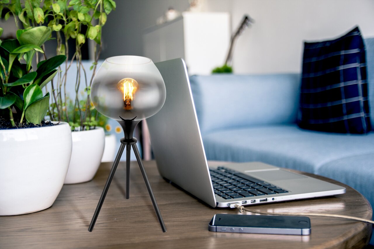 Moderne Tischleuchte mit dreibeinigem Stativ und kugelförmigem Glaslampenschirm, auf einem Tisch neben einem Laptop und Pflanzen platziert, seitliche Perspektive.