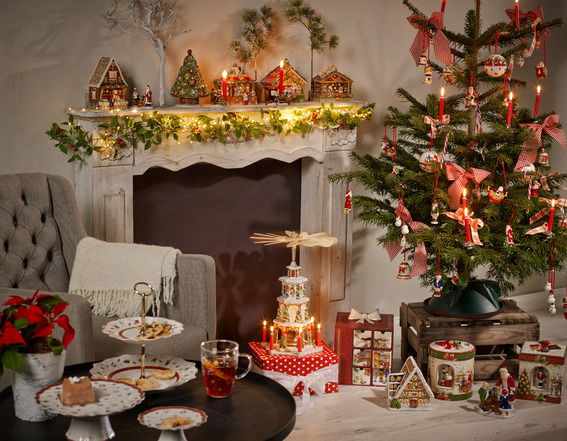 Gemütliche Weihnachtsdekoration mit Lebkuchenhaus und Spieluhr, im Vordergrund ein festlich gedeckter Tisch, im Hintergrund ein geschmückter Weihnachtsbaum und ein Kamin mit Lichtern, seitliche Perspektive.