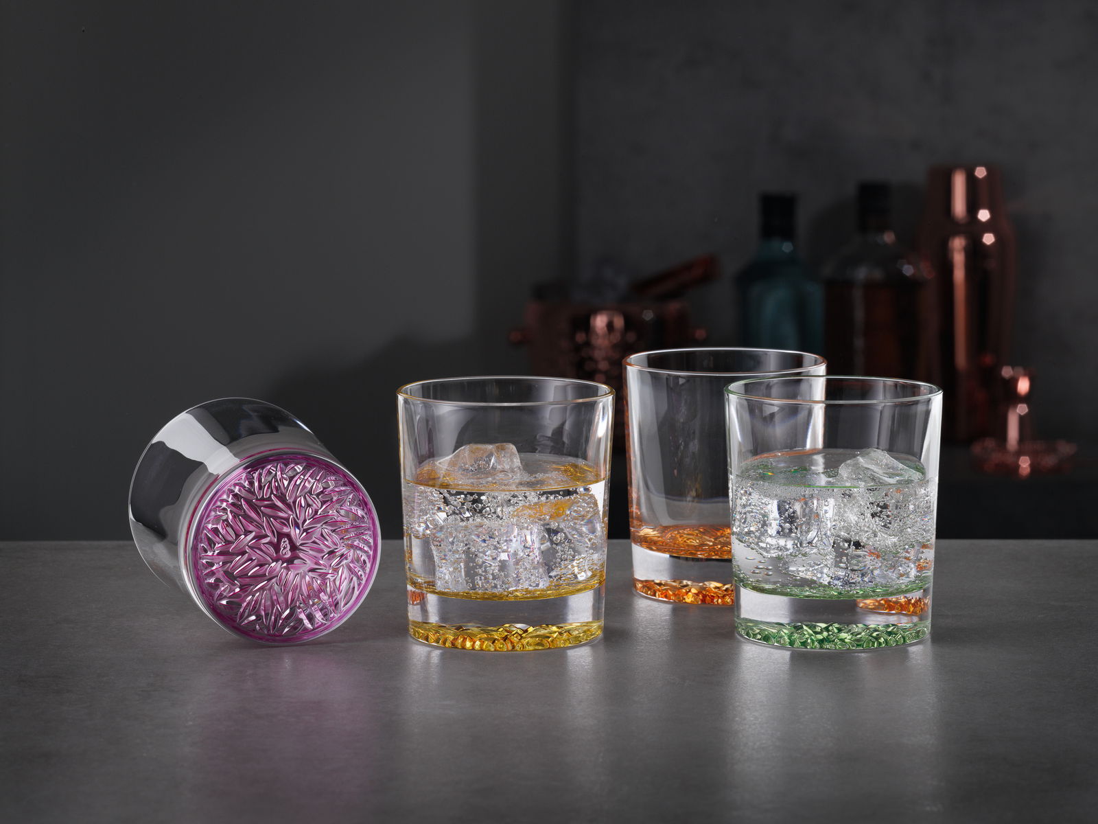 Vier Trinkbecher mit farbigen Böden in Lila, Gelb, Orange und Grün, gefüllt mit Eiswürfeln, auf einer dunklen Oberfläche. Perspektive: seitlich.