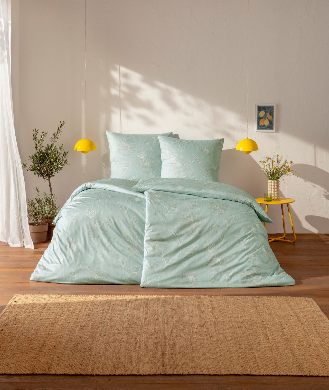 Estella Mako-Interlok-Jersey Bettwäsche 135/200cm EDESSA Mintgrüne Bettwäsche mit dezentem Blumenmuster auf einem Bett, frontal aus leicht erhöhter Perspektive fotografiert; daneben gelbe Pendelleuchten, gelber Beistelltisch mit Vase und Pflanze im hellen Schlafzimmer.