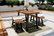 Moderner Gartentisch mit Holzplatte und schwarzen Metallbeinen, umgeben von gepolsterten Bänken, auf einer Terrasse mit Pflanzen im Hintergrund. Perspektive von schräg vorne.