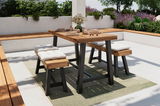 Moderner Gartentisch mit Holzplatte und schwarzen Metallbeinen, umgeben von gepolsterten Bänken, auf einer Terrasse mit Pflanzen im Hintergrund. Perspektive von schräg vorne.