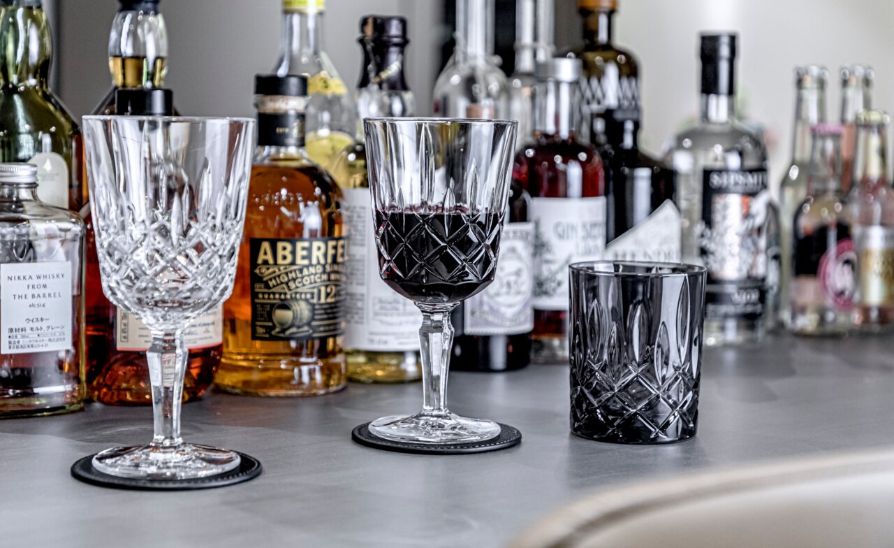 Nachtmann Cocktailset 2-tlg. NOBLESSE Drei elegante Kristallgläser in Nahaufnahme, umgeben von verschiedenen Spirituosenflaschen auf einer Bar.