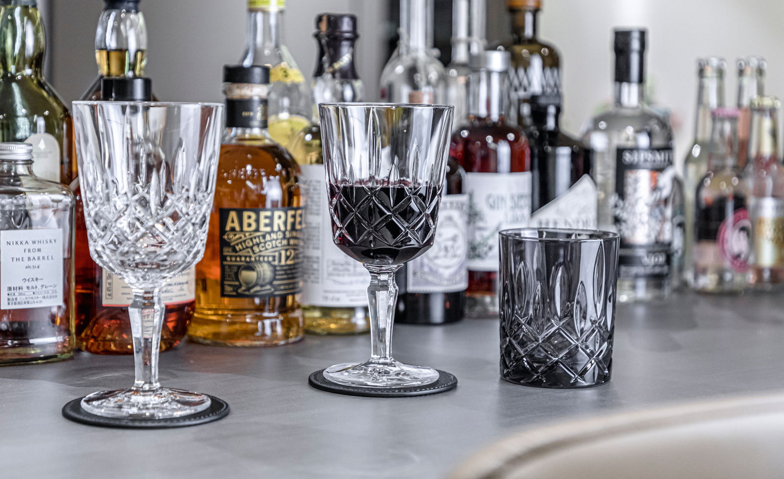 Nachtmann Cocktailset 2-tlg. NOBLESSE Drei elegante Kristallgläser in Nahaufnahme, umgeben von verschiedenen Spirituosenflaschen auf einer Bar.
