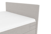 Gepolstertes Boxspringbett mit hohem, rechteckigem Kopfteil in hellgrauem Stoffbezug, Detailansicht in schräger Frontperspektive von der Seite