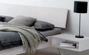 Modernes Bettgestell mit grauer Bettwäsche und Kissen, seitliche Perspektive, daneben ein Nachttisch mit Lampe und Büchern.