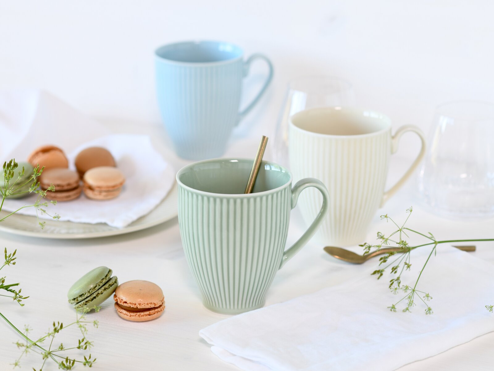 Pastellfarbene, geriffelte Keramikbecher mit Henkel auf hellem Tisch, vorne mintgrüner Becher mit Löffel, im Hintergrund blaue und cremefarbene Tasse, dazu Macarons und zarte Blumen, aufgenommen in leichter Schrägansicht von vorn.