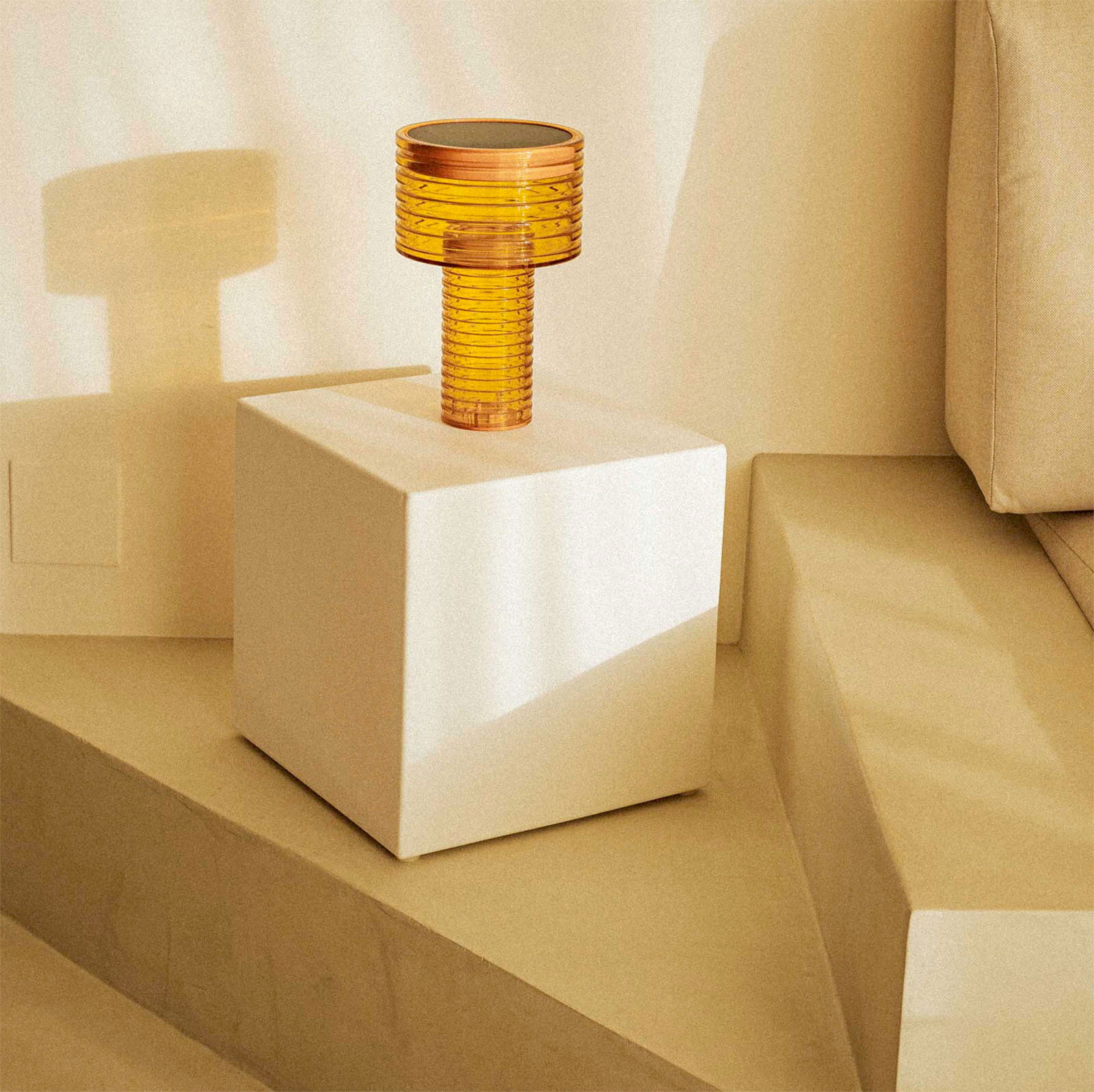 Bernsteinfarbene, geriffelte Tischlampe aus Glas auf weißem Würfelpodest neben beigem Sofa, in warmem Licht, aus leicht erhöhter Frontansicht.