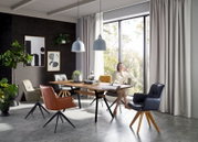 Schuine vooraanzicht van een moderne eetkamer met houten eettafel en vier kuipstoelen met draaivoet in verschillende kleuren, bij grote ramen met gordijnen en hanglampen.