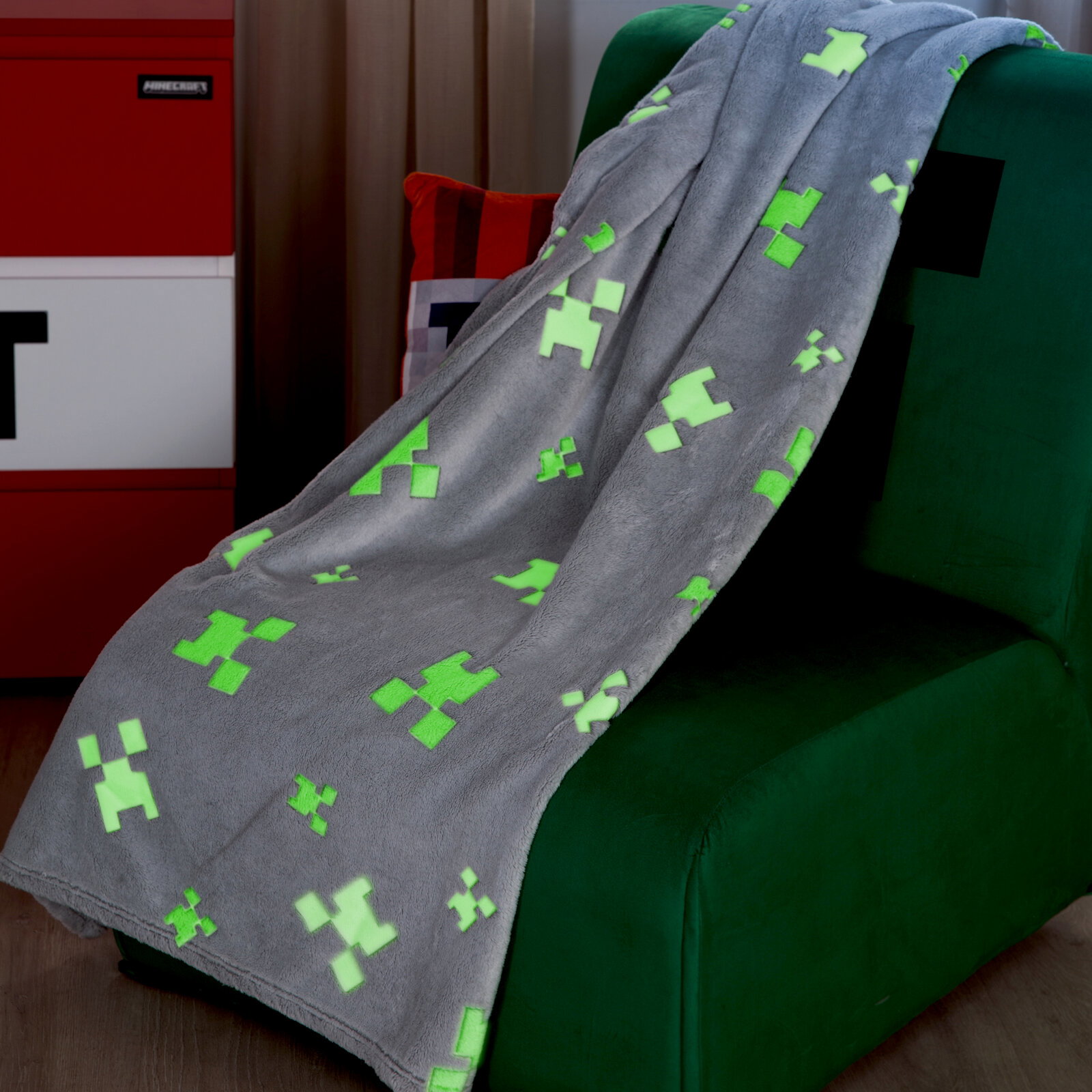 Graue, flauschige Minecraft-Creeper-Decke mit leuchtend grünen Pixel-Motiven, über einen grünen Sessel drapiert, seitliche Perspektive