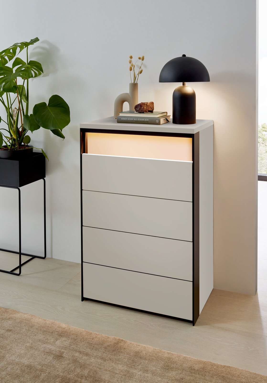 Voleo Hochkommode ZAYA Hochkommode mit vier Schubladen, dekoriert mit einer Lampe und Büchern, aus einer seitlichen Perspektive fotografiert.
