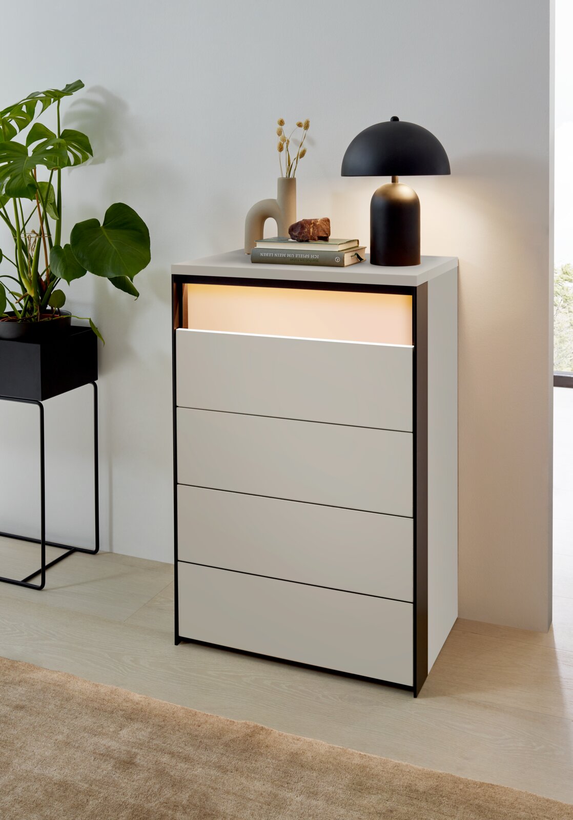 Voleo Hochkommode ZAYA Hochkommode mit vier Schubladen, dekoriert mit einer Lampe und Büchern, aus einer seitlichen Perspektive fotografiert.