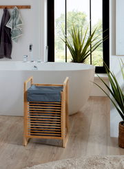 Badezimmer-Szene mit freistehender weißer Badewanne und modernem Wasserhahn; im Vordergrund ein kleiner Bambus-Wäschekorb mit dunkelblauem Stoffeinsatz, frontal aus leicht erhöhter Perspektive fotografiert.