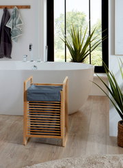 Badezimmer-Szene mit freistehender weißer Badewanne und modernem Wasserhahn; im Vordergrund ein kleiner Bambus-Wäschekorb mit dunkelblauem Stoffeinsatz, frontal aus leicht erhöhter Perspektive fotografiert.