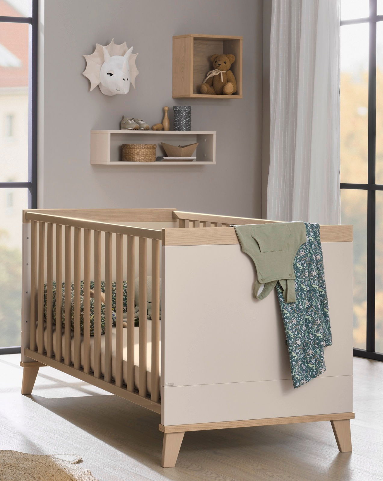 Kinderzimmer mit einem modernen, hellen Gitterbett aus Holz, das von der Seite gezeigt wird. An der Wand hängt eine quadratische Wandbox mit einem Teddybär.