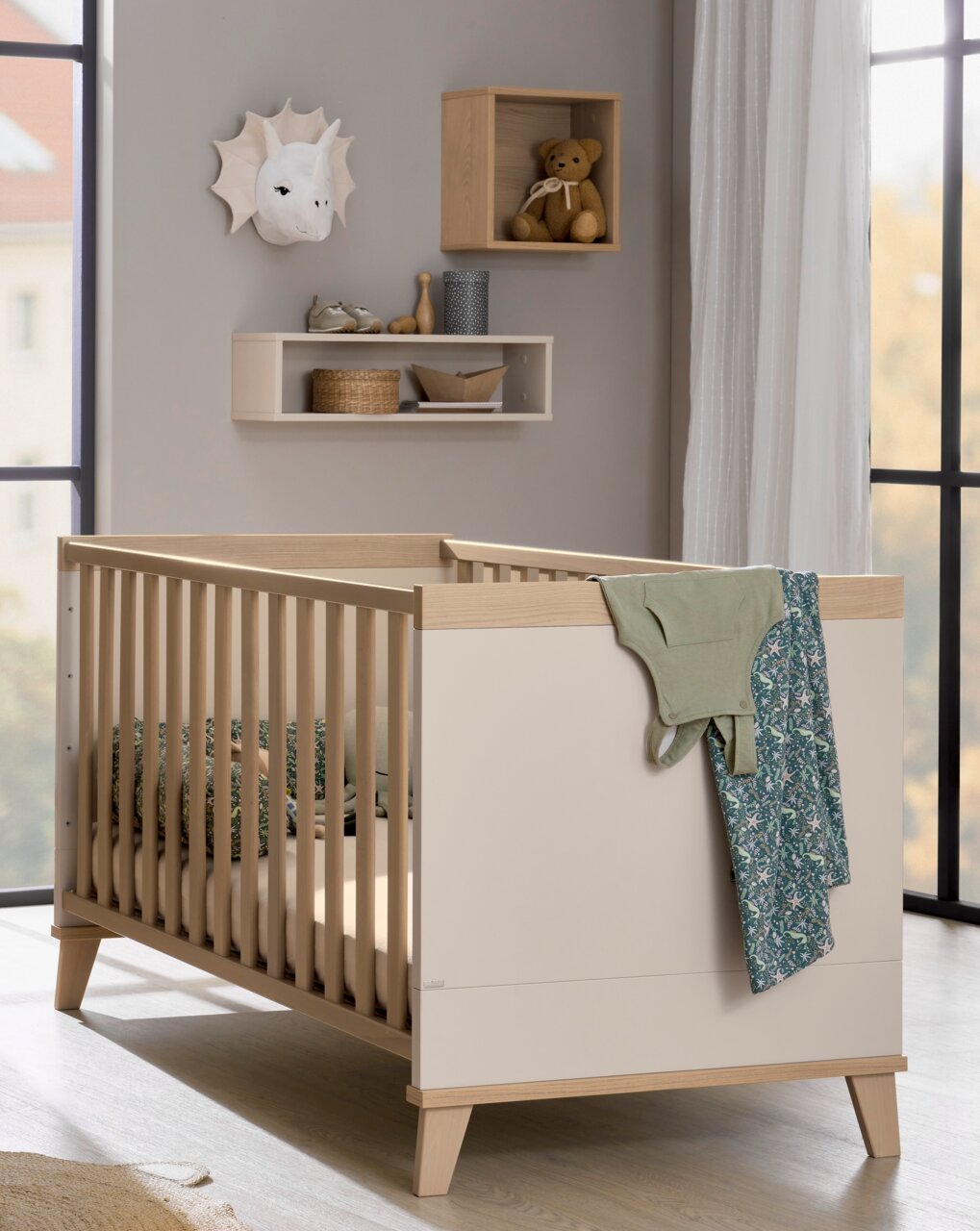 Kinderkamer met een modern, licht houten ledikantje, dat vanaf de zijkant te zien is. Aan de muur hangt een vierkant wandrekje met een teddybeer.