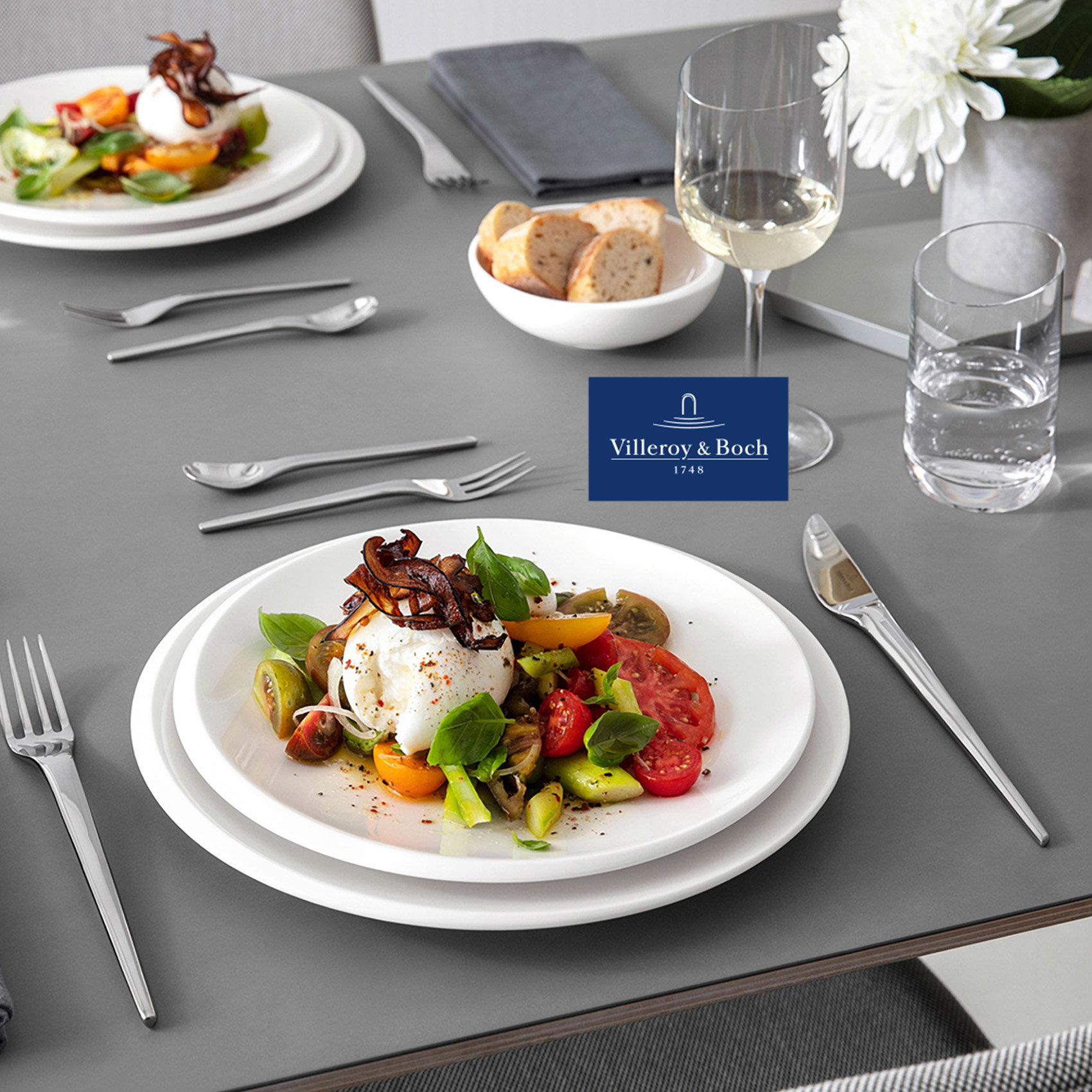 Modern gedeckter Esstisch mit weißem Villeroy & Boch Geschirr, Edelstahlbesteck, Weißweinglas und Burrata-Tomatensalat auf grauer Tischplatte.