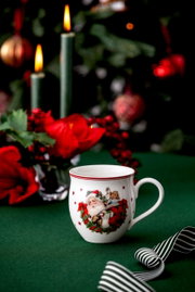 Weihnachtlicher Henkelbecher mit Santa Claus-Motiv, umgeben von festlicher Dekoration, aus seitlicher Perspektive fotografiert.
